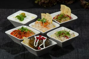 A vibrant assortment of Middle Eastern mezze dishes in white bowls on dark fabric.